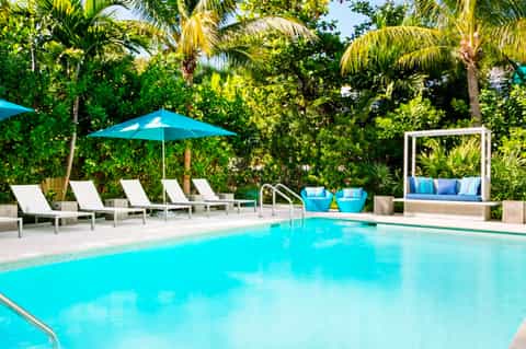 Turquoise swimming pool with white lounge chairs, blue umbrella, and tropical palm trees in background