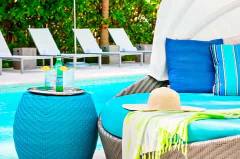 Luxurious poolside lounge with turquoise woven ottoman table, blue cushioned seating, and tropical greenery backdrop