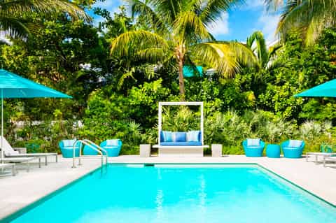 Turquoise rectangular pool surrounded by lush tropical palms, white seating areas, and teal umbrellas