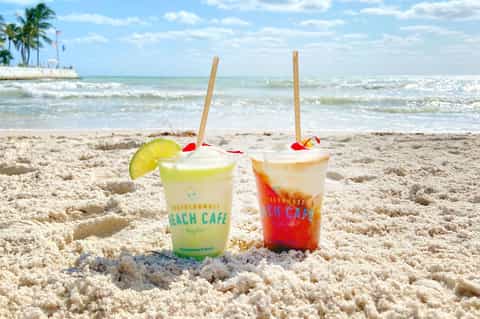 Two colorful beverages from Southernmost Beach Cafe sitting in sand with ocean and palm trees in background
