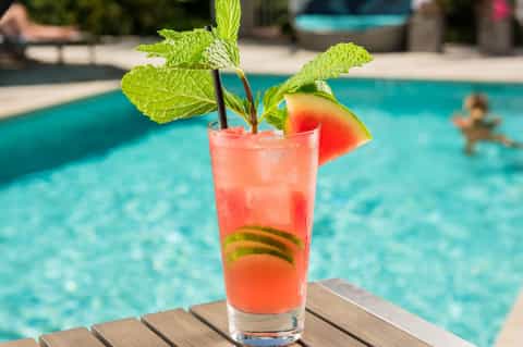 Tropical cocktail with watermelon garnish and fresh mint on poolside wooden table with swimmers in background