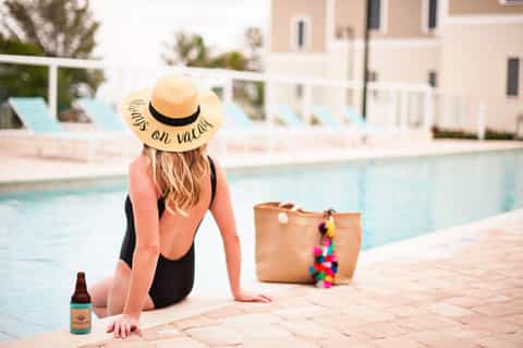 Woman in swimsuit and straw hat sitting poolside with beach bag and beverage bottle
