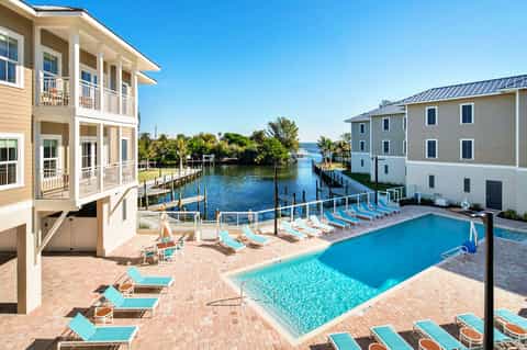 Waterfront resort with pool, dock, and multi-story residential buildings surrounded by palm trees and blue sky