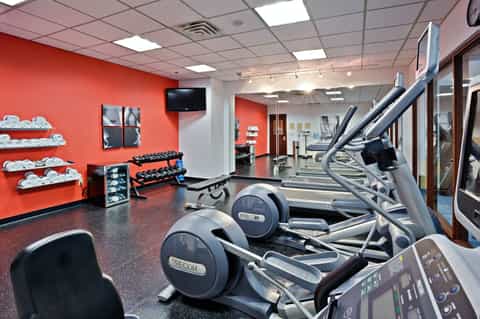 Modern gym facility with red accent walls, cardio equipment, dumbbells on racks, and workout machines