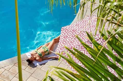 Person sunbathing on pink patterned towel beside bright blue pool with green plants