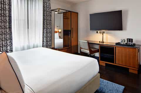 Modern hotel bedroom with white bedding, wooden wardrobe, desk with TV, and patterned curtains
