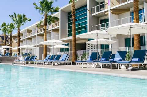 Modern resort pool with palm trees, white umbrellas, and blue loungers beside contemporary building