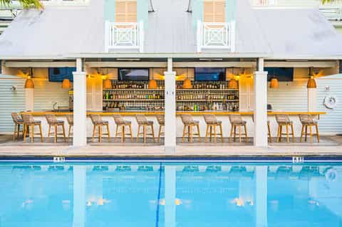 Modern poolside bar with white columns, wooden bar stools, extensive liquor selection, and clear pool water