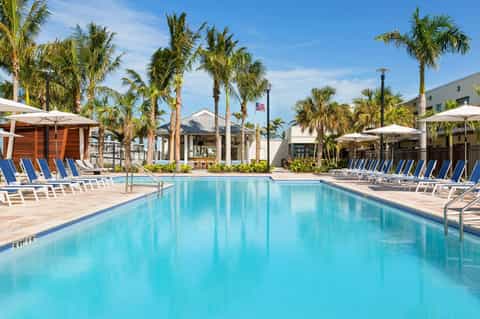 Large resort pool with turquoise water, palm trees, blue loungers, and white umbrellas under clear sky