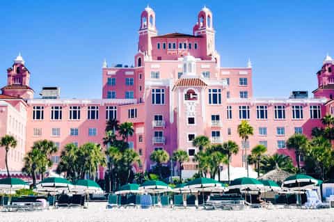Pink historic beachfront hotel with twin towers, palm trees, and beach loungers