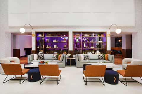 Modern hotel lobby with purple-lit shelving, gray sofas, orange accent chairs, and curved arc lamps