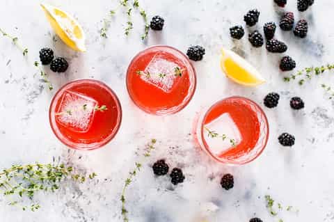 Three red berry cocktails with strawberry garnish, surrounded by blackberries and lemon slices on marble