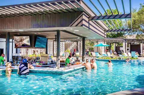 Resort pool with swim-up bar area, modern pergola shade structure, and guests enjoying water activities