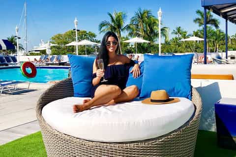 Woman relaxing in woven lounge chair with blue pillows by resort pool with palm trees and boats