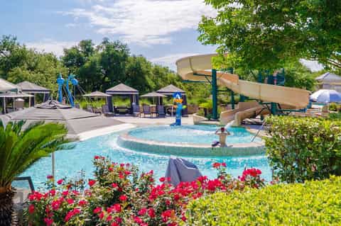 Resort pool area with water slides, cabanas, loungers, flowering plants, and lush green landscaping