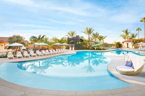 Large resort pool with water slide, lounge chairs under blue umbrellas, and palm trees surrounding the area