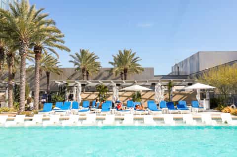 Resort pool deck with bright blue loungers, white umbrellas, palm trees, and modern buildings
