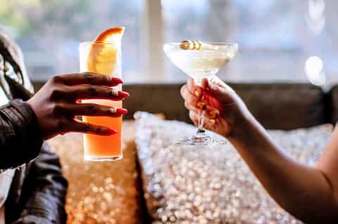 Two people toasting with cocktails at sunset, holding colorful drinks with citrus garnishes
