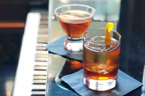 Two cocktails on a piano keyboard - amber drink with lemon twist and dark cocktail with foam top