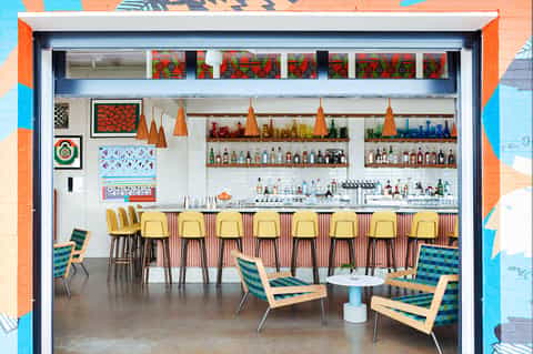 Colorful bar interior with yellow stools, shelves of liquor bottles, and vibrant artwork on exposed brick walls