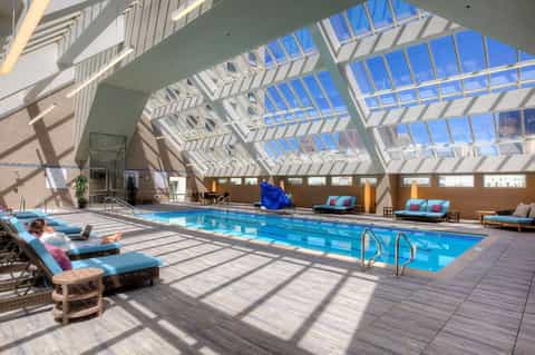 Indoor swimming pool with skylights, lounge chairs, and blue water under a modern glass ceiling