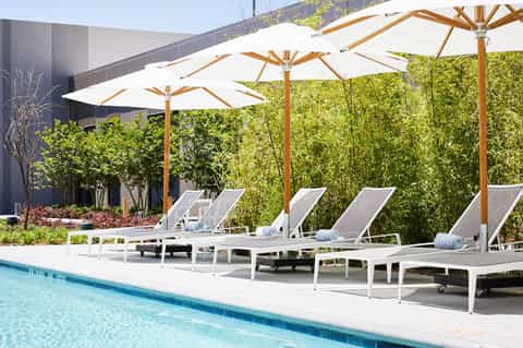 Modern poolside lounge area with white chaise lounges under wooden-frame white umbrellas and green privacy hedges