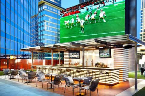 Outdoor bar with striped design and seating, large sports display screen above, modern urban setting with tall buildings