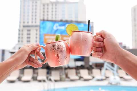 Two hands holding pink cocktails with citrus garnish against urban rooftop pool backdrop
