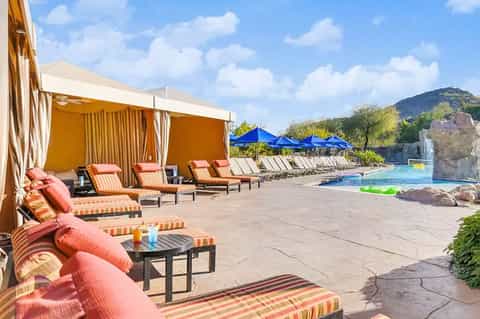 Luxurious poolside area with orange cabanas, striped loungers, blue umbrellas, and mountain views
