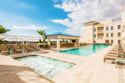 Resort pool area with spa tub, cabana, tan loungers, and multi-story yellow building under blue sky