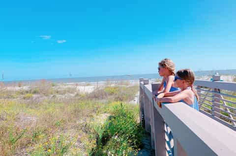 Two children on wooden beach boardwalk overlook dunes and ocean under clear blue sky