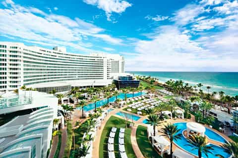 Beachfront resort with high-rise hotel, multiple pools, palm trees, and ocean view under blue sky