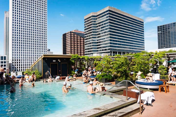 Rooftop pool at urban hotel with skyline views, guests swimming, lounge areas, and greenery