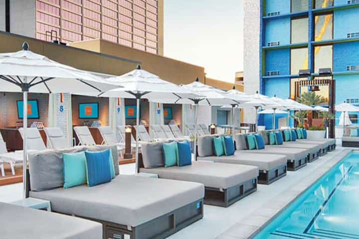 Resort pool deck with white umbrellas, gray lounge chairs, colorful pillows, and turquoise water