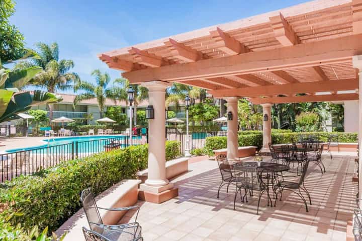 Covered patio with red-brown pergola overlooking resort pool and palm tree landscaping