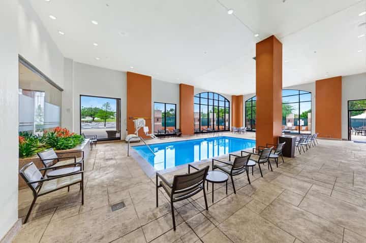 Indoor resort pool with blue water, orange accent columns, lounge chairs, and large windows overlooking outdoor areas