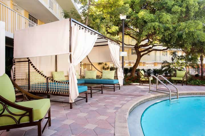 Pool courtyard with white cabanas, lounge seating, mature tree, and resort buildings