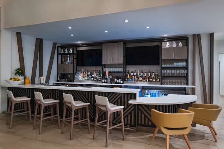 Modern hotel bar with dark geometric wood design, white bar seating, and liquor display