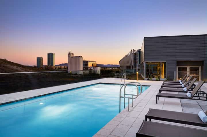 Rooftop pool at twilight with loungers, city skyline backdrop, architectural building, and illuminated water