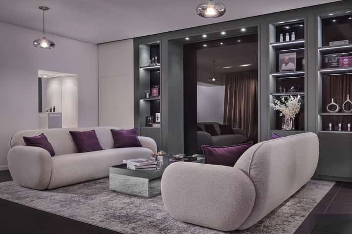Luxury spa lounge with curved beige seating, dark shelving with beauty products, and soft pendant lighting