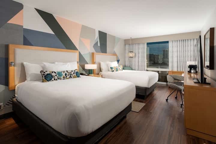 Modern hotel bedroom with two queen beds, geometric wall art, desk, and city view windows