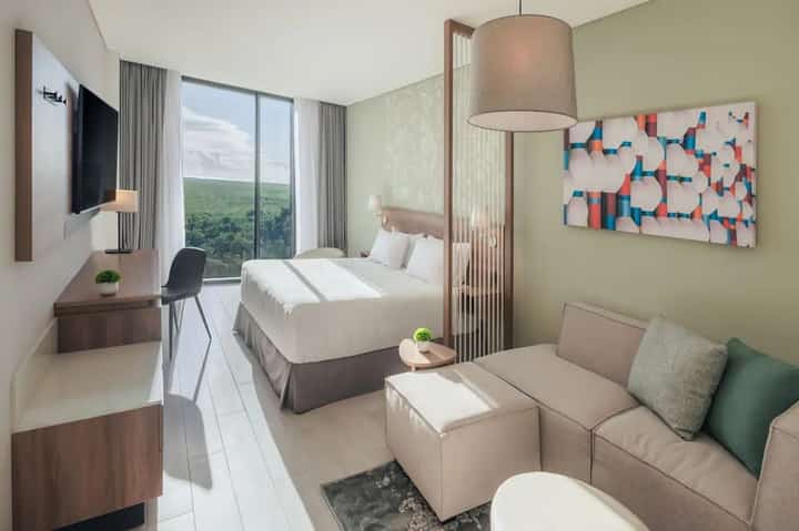 Hotel room with bed, sofa, work desk, TV, and large window overlooking green landscape
