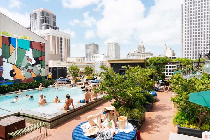 Urban rooftop pool with colorful art mural, swimmers, lush greenery, and city skyscraper views
