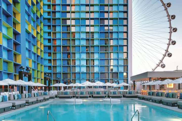 Resort pool deck with colorful building facade, white umbrellas, and London Eye visible