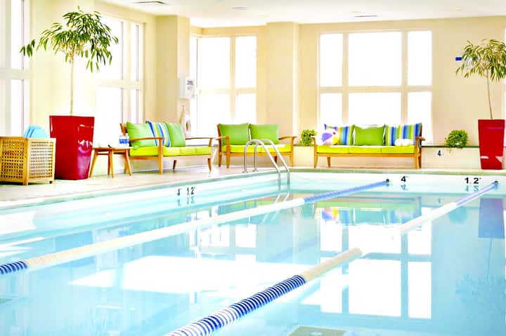 Indoor lap pool with lane dividers, wooden lounge chairs with colorful pillows, large windows, and potted plants