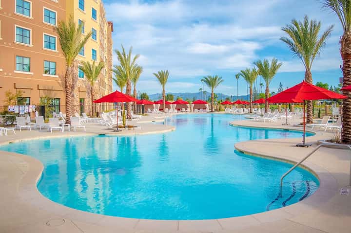 Large lagoon-style resort pool with red umbrellas, palm trees, and sunny mountain backdrop