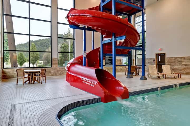 Indoor aquatic center with red spiral waterslide and turquoise pool, mountain views through windows