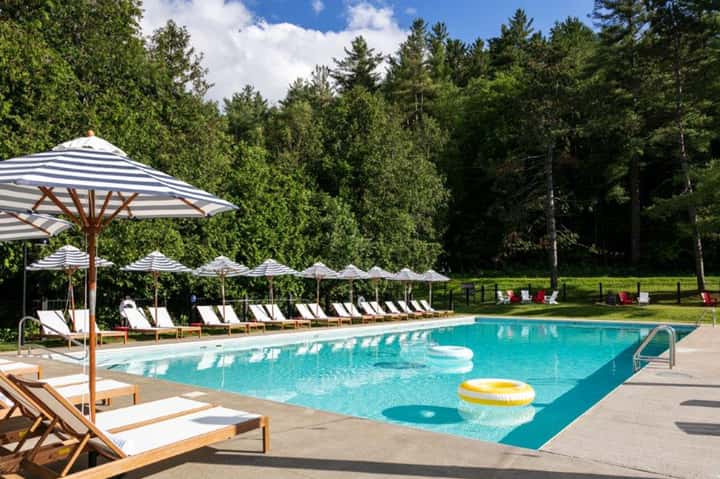 Resort pool surrounded by white lounge chairs, striped umbrellas, and coniferous forest landscape
