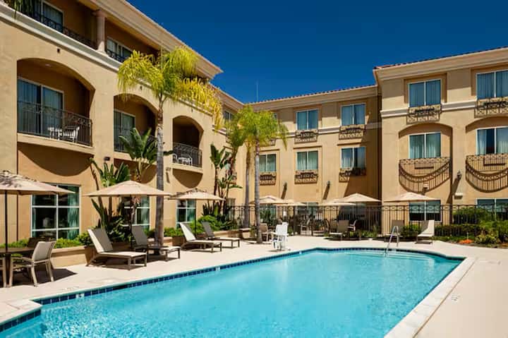 Resort courtyard with rectangular pool, tan buildings with arches, and palm trees under clear blue sky