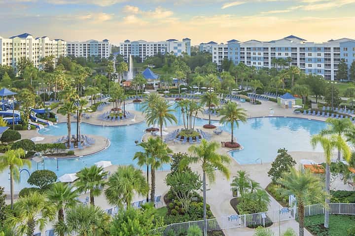 Resort pool complex with multiple pools, palm trees, lounge chairs, and multi-story residential buildings